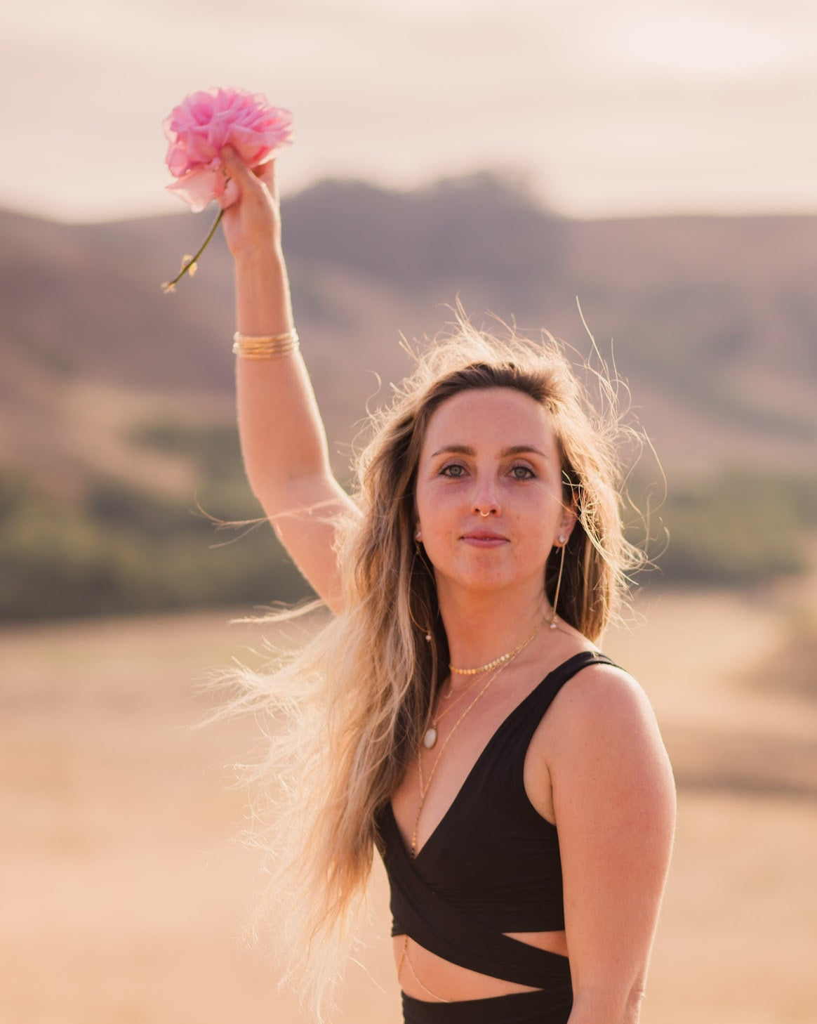 Woman in a black outfit holding a pink flower in an outdoor setting with mountains in the background