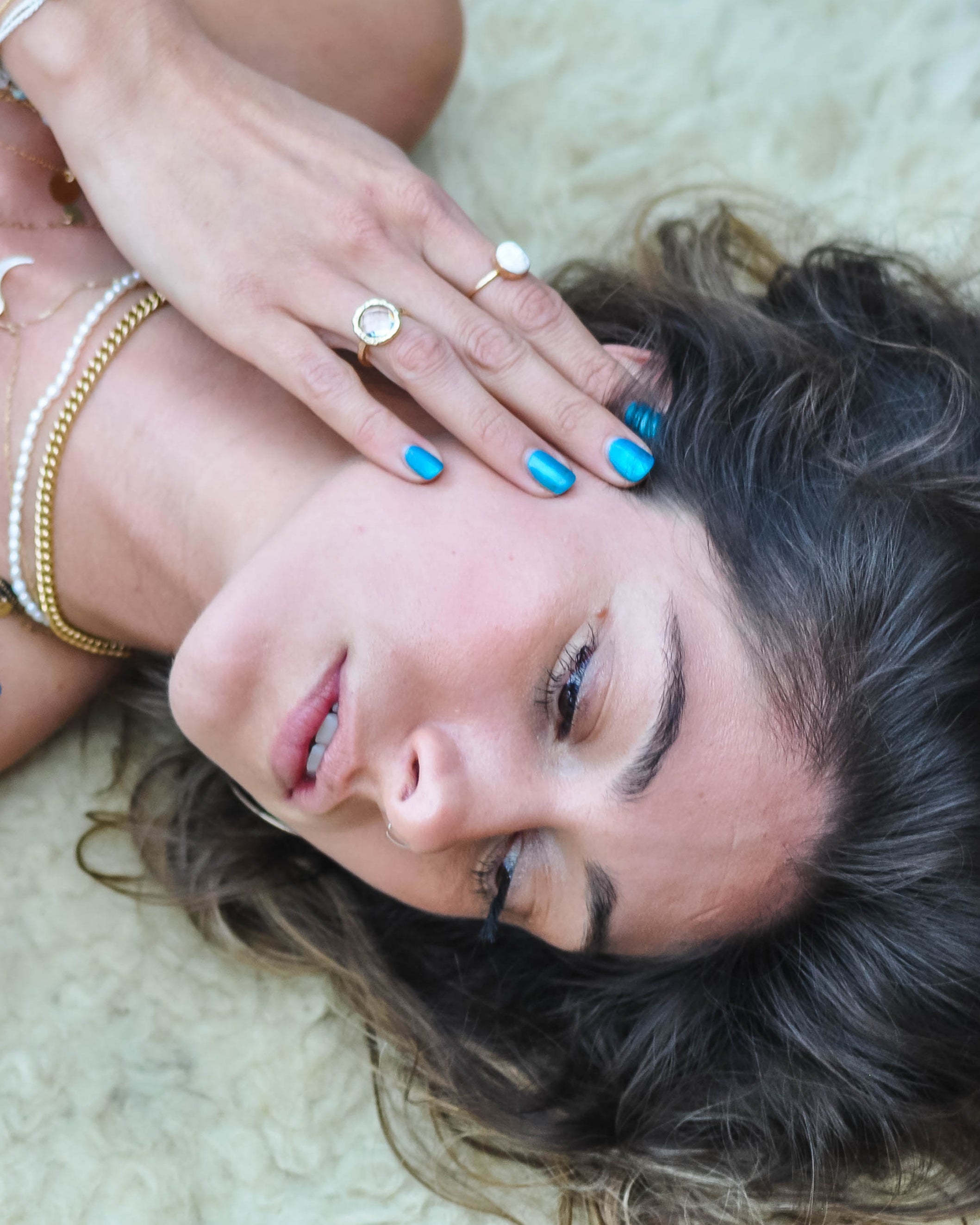 Woman lying on a light-colored surface with jewelry and blue nail polish.
