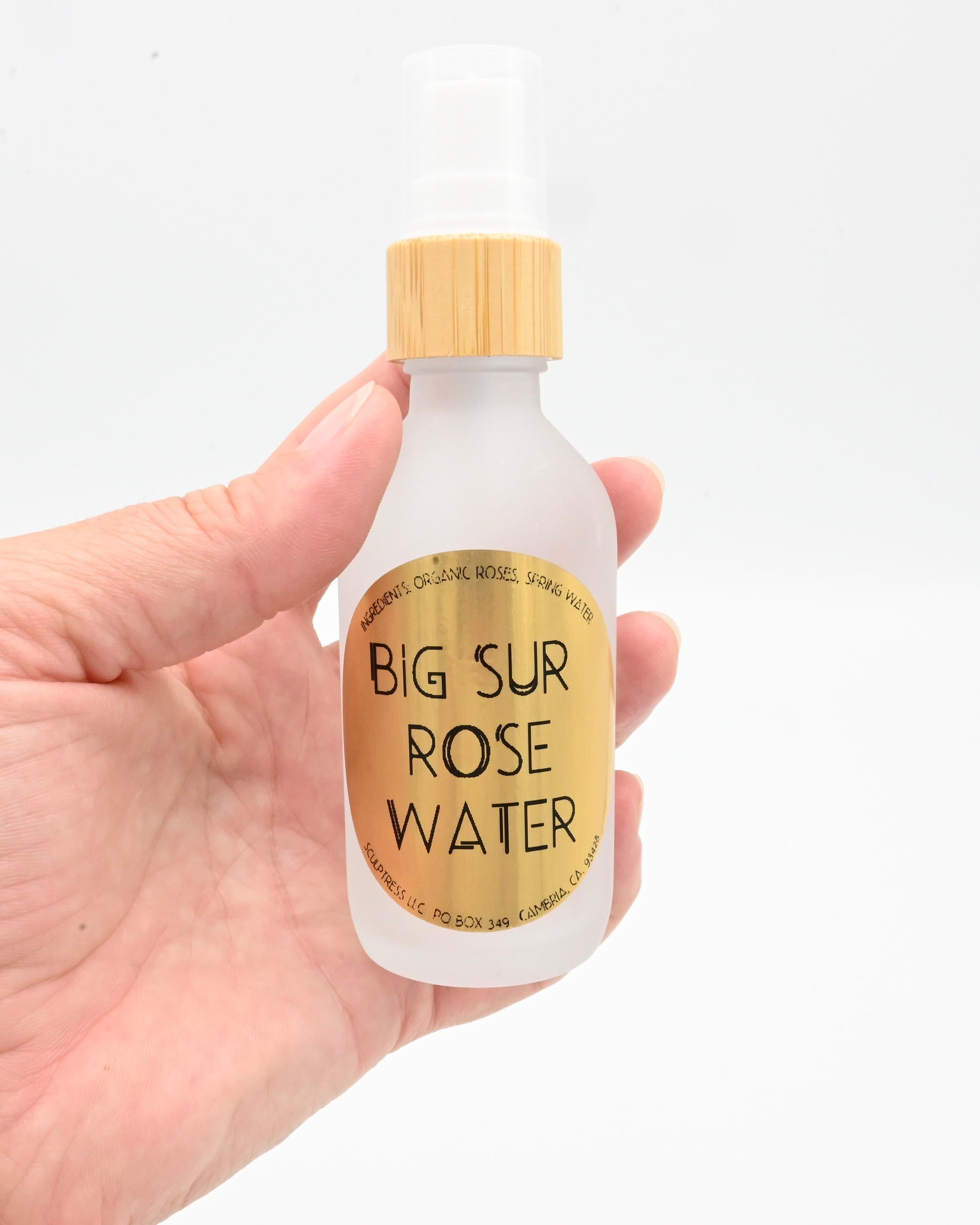 Hand holding a bottle of 'Big Sur Rose Water' with a gold cap on a white background