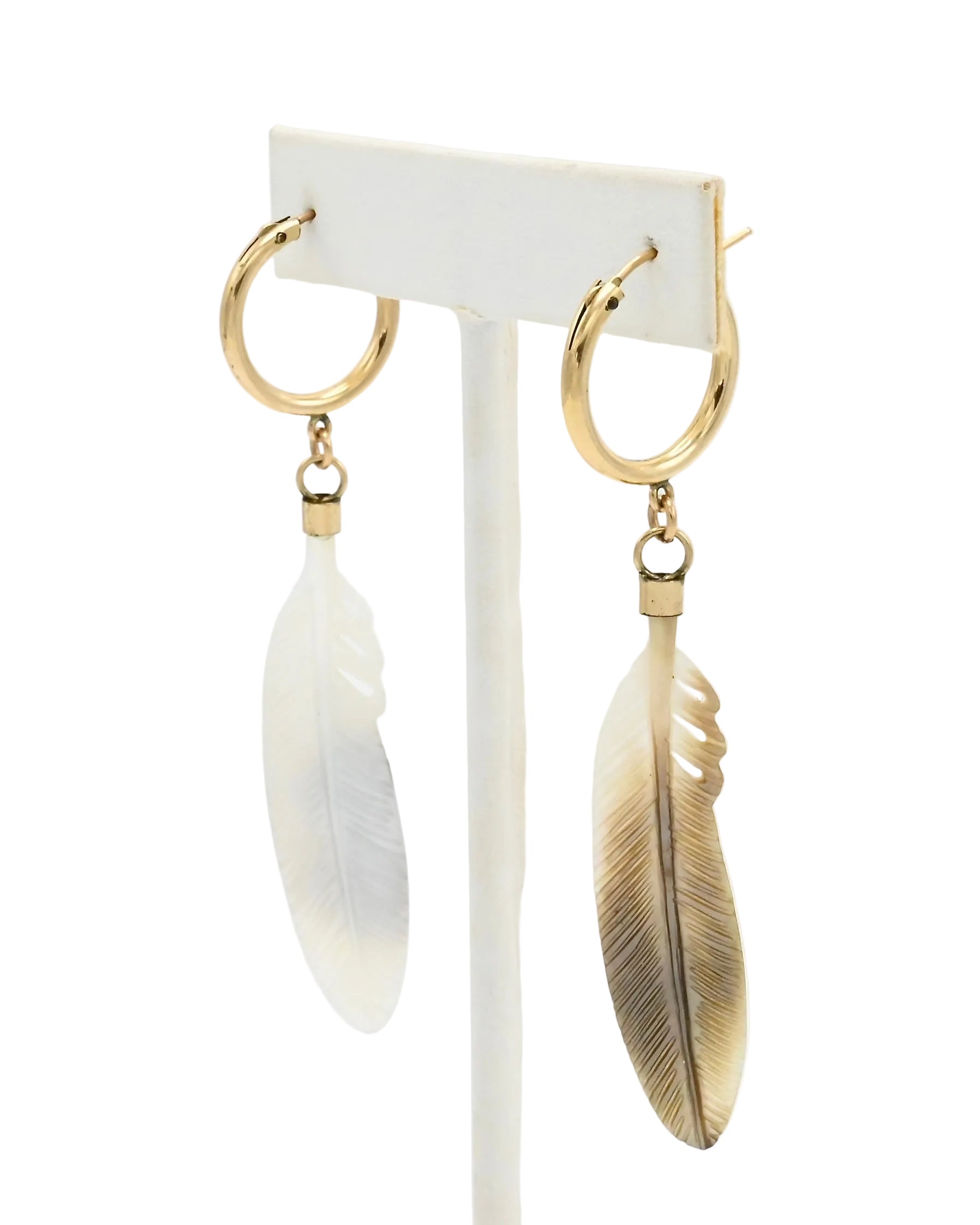 A pair of gold hoop earrings with dangling feather charms, displayed on a white stand against a white background. One feather is white, and the other is a warm champagne-gold tone.