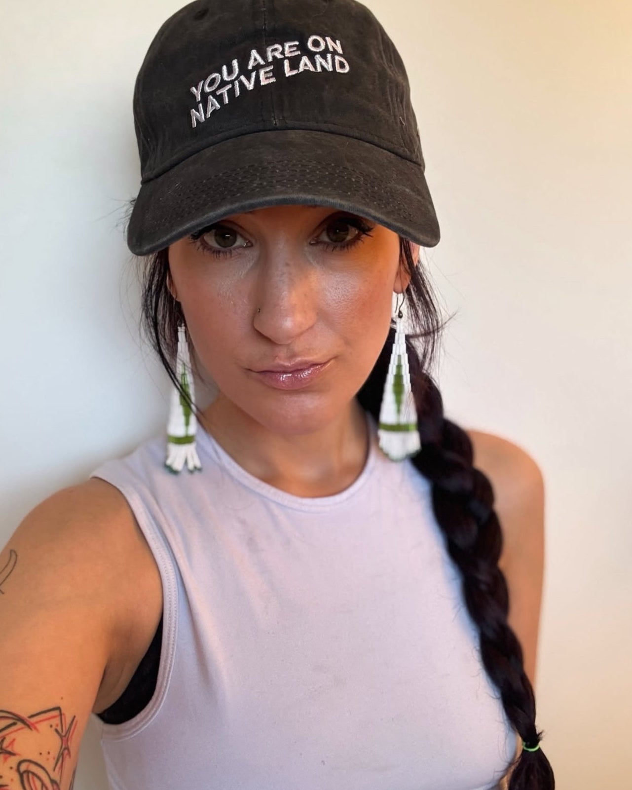 Person wearing a black cap with 'YOU ARE ON NATIVE LAND' text, white tank top, and braided hair against a plain background.